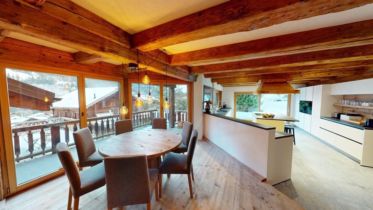 Rustic dining room with wooden beams and a round table, overlooking a snow-covered balcony with mountain views.