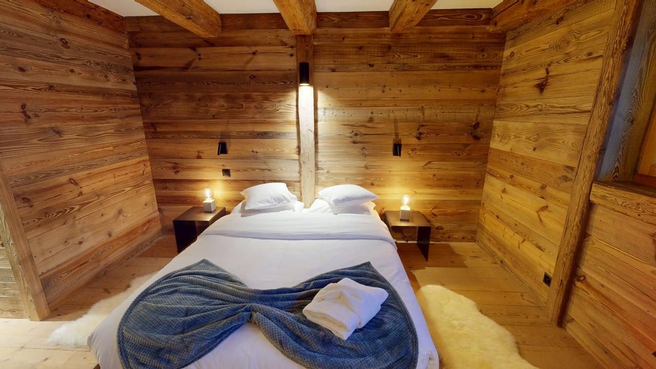 A cozy bedroom with wood-paneled walls, a bed with white linens and a blue textured throw, and warm bedside lighting.