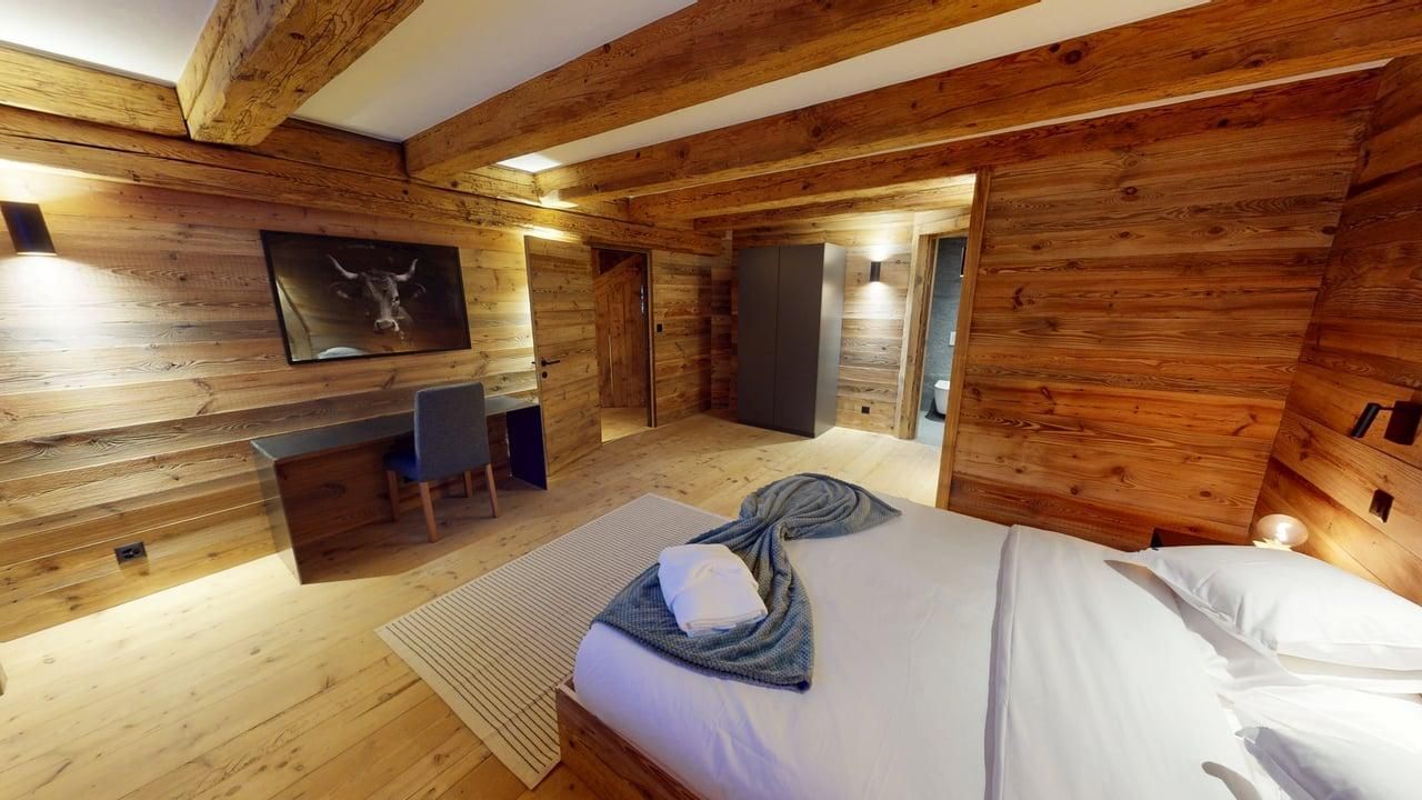 A rustic bedroom with wooden walls and ceiling, featuring a bed with white linens, a desk, and a chair.