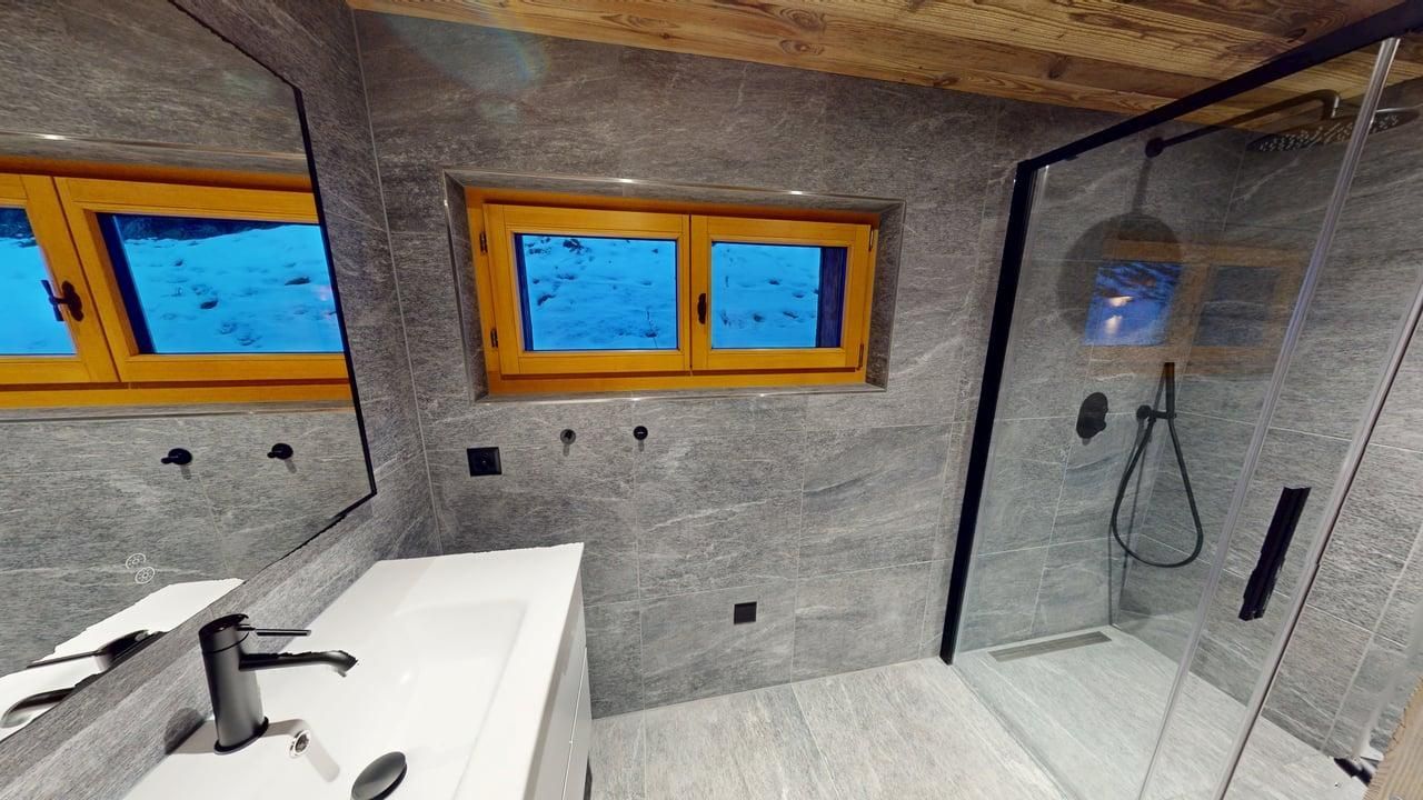 A modern bathroom with grey stone-tiled walls, a white vanity sink, and a walk-in glass shower beneath a wooden ceiling.