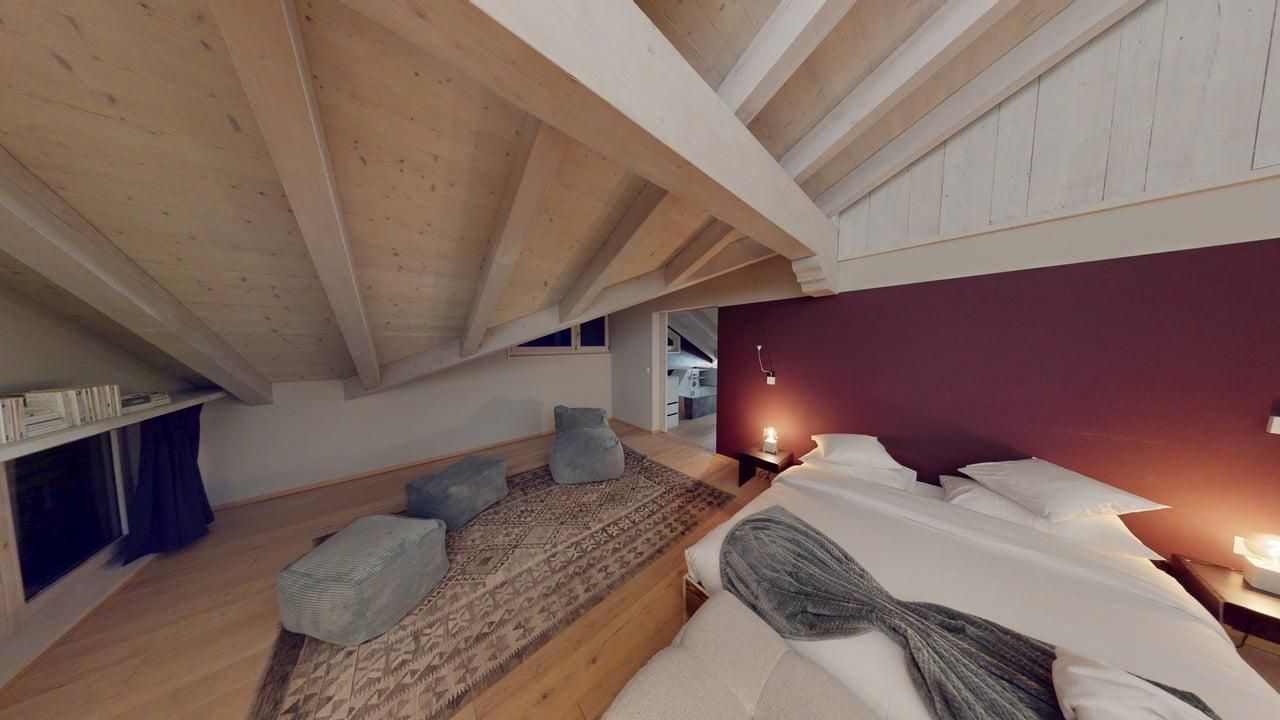 An attic bedroom with a sloped wooden ceiling, a large bed with white linens, and grey floor cushions on a textured rug.