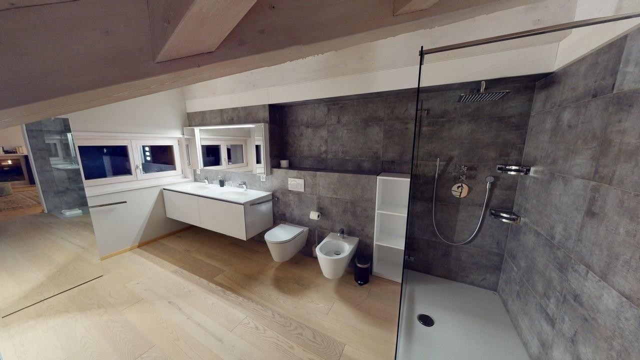 A modern bathroom in an attic with gray tiled walls, a white vanity, a toilet, a bidet, and a walk-in shower.