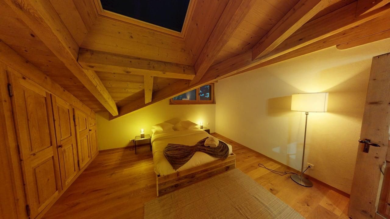 An attic bedroom with warm lighting, a wooden bed, sloped ceiling beams, a floor lamp, and built-in wooden closets.