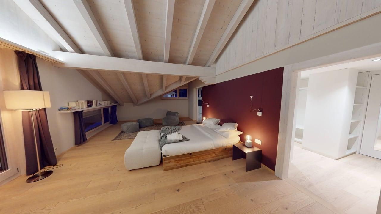 A spacious bedroom with light wood flooring, a white bed with a wooden frame, a maroon accent wall, and a sloped ceiling.