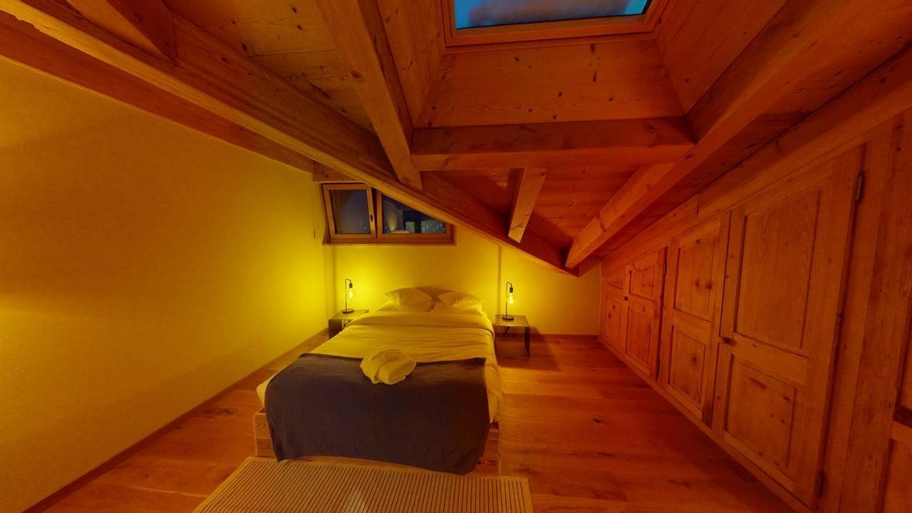 A cozy attic bedroom with wooden beams and ceiling, featuring a bed, warm lighting, and a skylight.