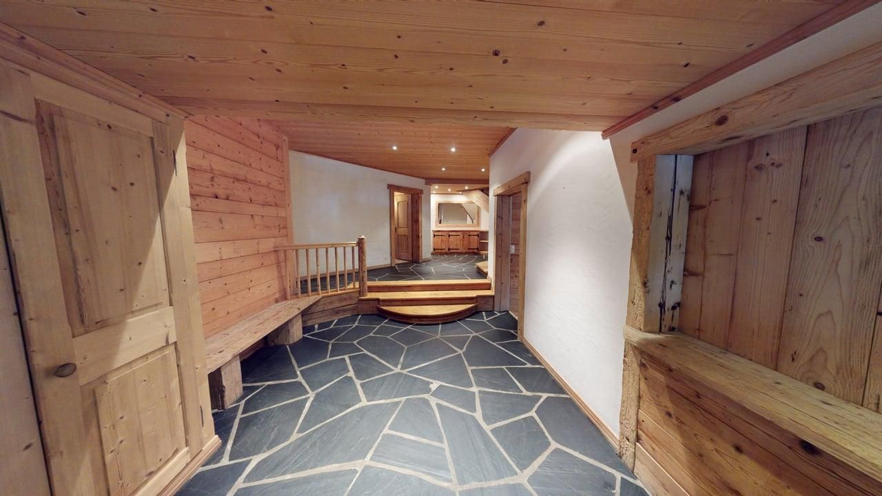 A rustic hallway with stone flagstone flooring, wooden walls, and a small set of wooden stairs leading to a lighted room.