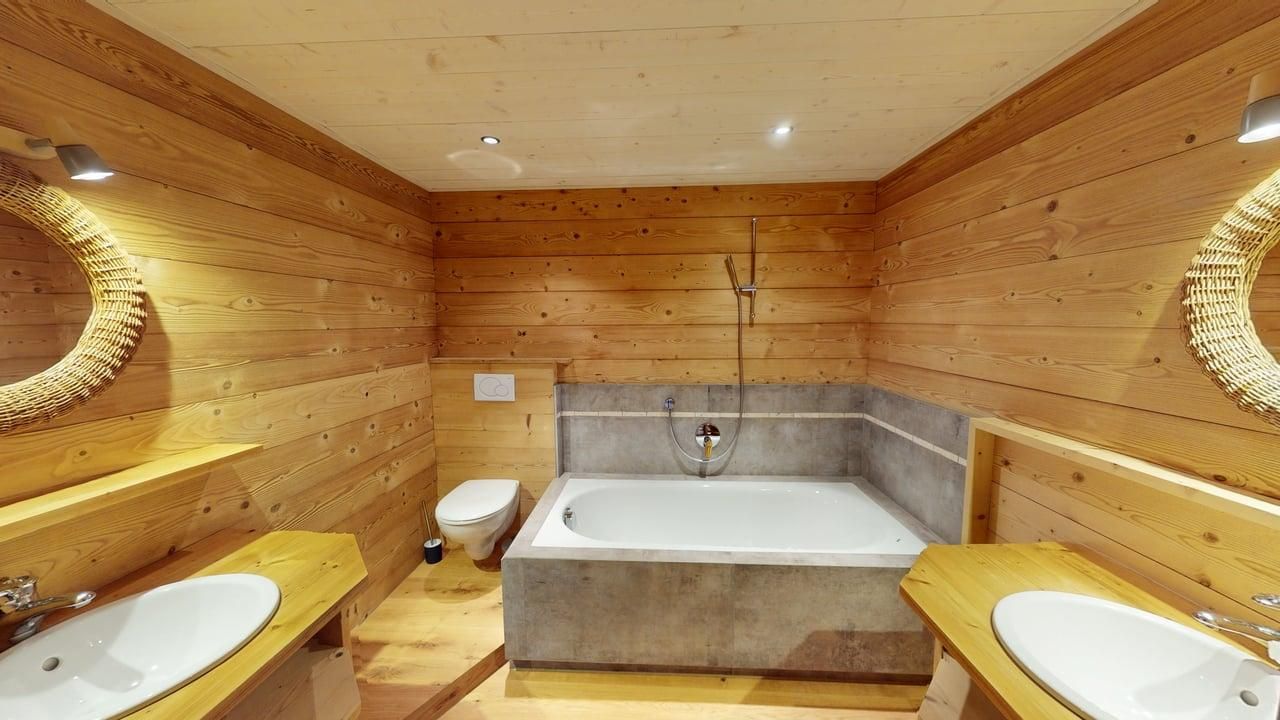 A modern bathroom with wood-paneled walls, a sunken tub with concrete surround, two pedestal sinks, and a wall-hung toilet.