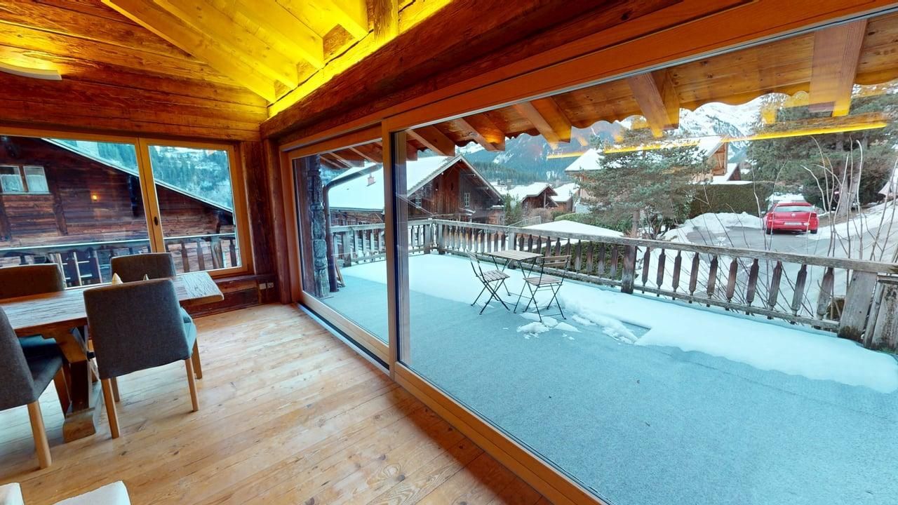 A cozy, wooden indoor dining area with large windows overlooking a snow-covered balcony and a mountainous winter landscape.