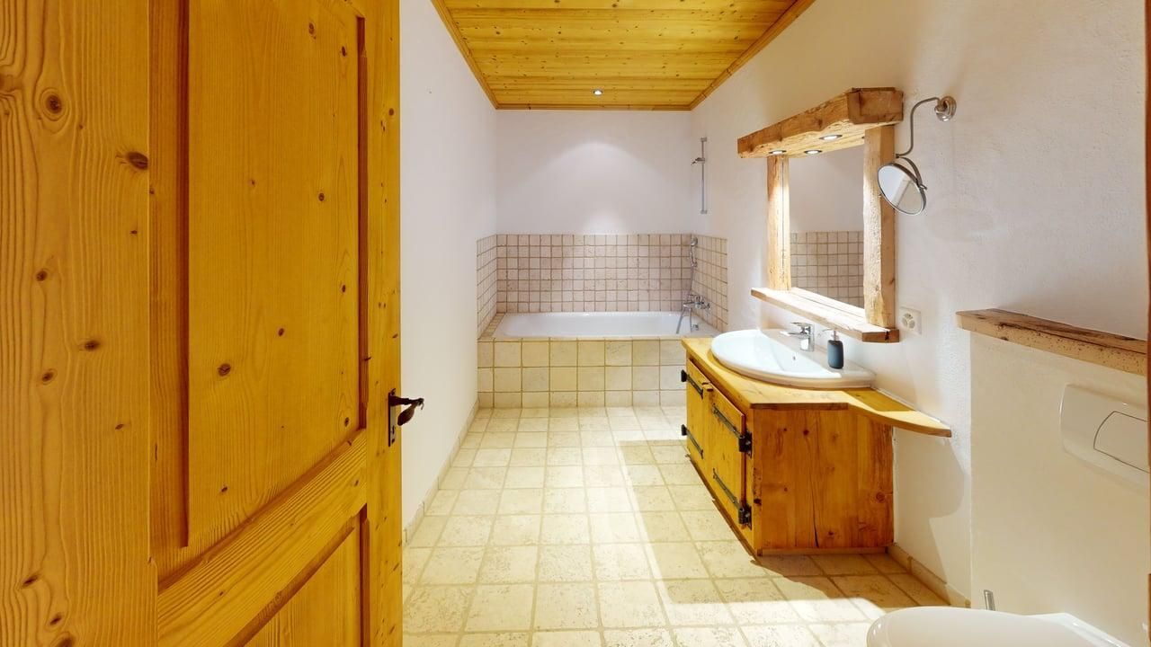 A bathroom with a wooden door, tiled floor, a vanity with a sink and mirror, and a bathtub at the back.
