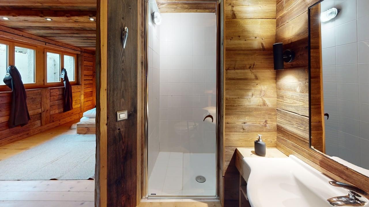 A bathroom with rustic wood walls, a glass-enclosed shower, a white sink, and a view into a wooden bedroom.