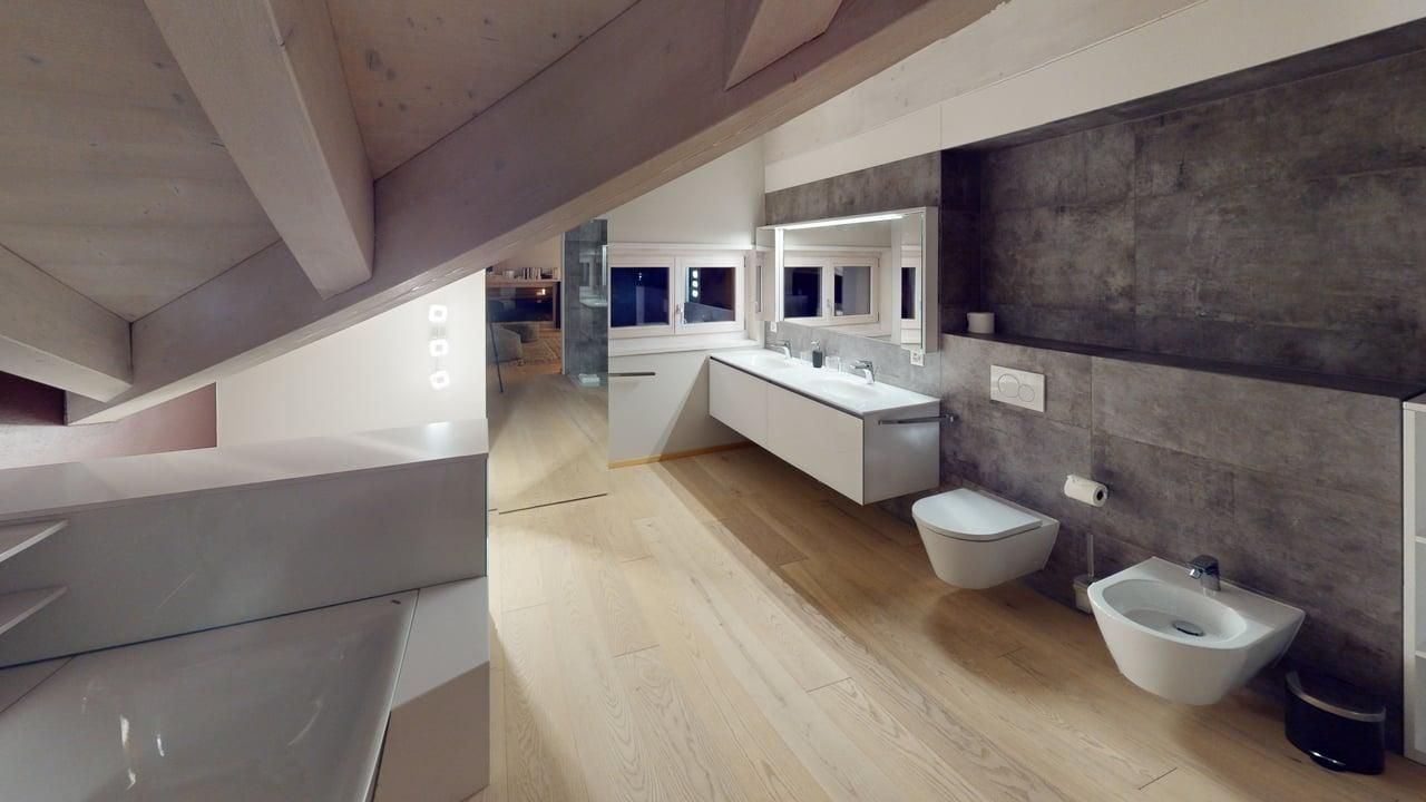 A modern, minimalist bathroom with a sloped ceiling, wood flooring, a white floating vanity, and charcoal-tiled walls.