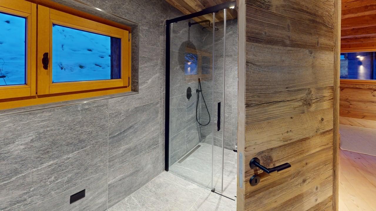 Modern bathroom with gray stone walls, a glass-enclosed shower, a wood-framed window, and a rustic wooden door.