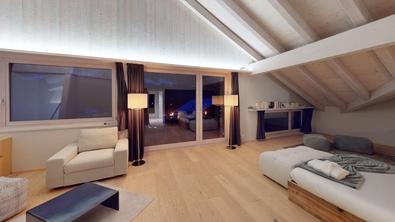Modern attic hotel room with light wood floors, a white armchair, a bed with gray bedding, and floor-to-ceiling windows.