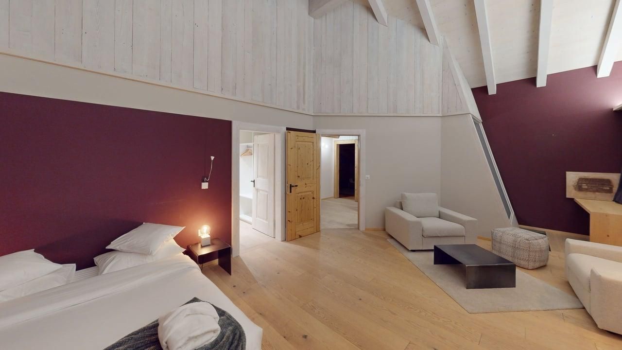 A modern hotel room with a bed, seating area, wooden floors, and a vaulted white ceiling against dark purple walls.