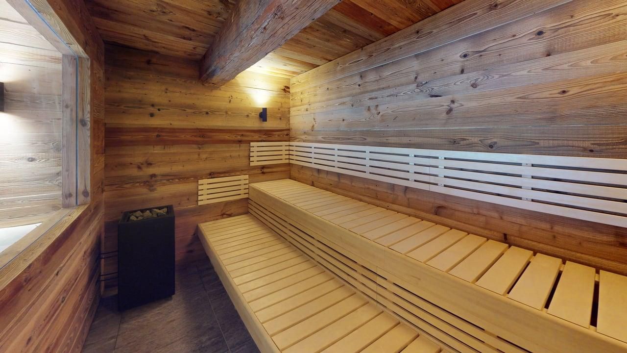 A wooden sauna room featuring tiered light-wood benches and a black heater against a rustic paneled wall.