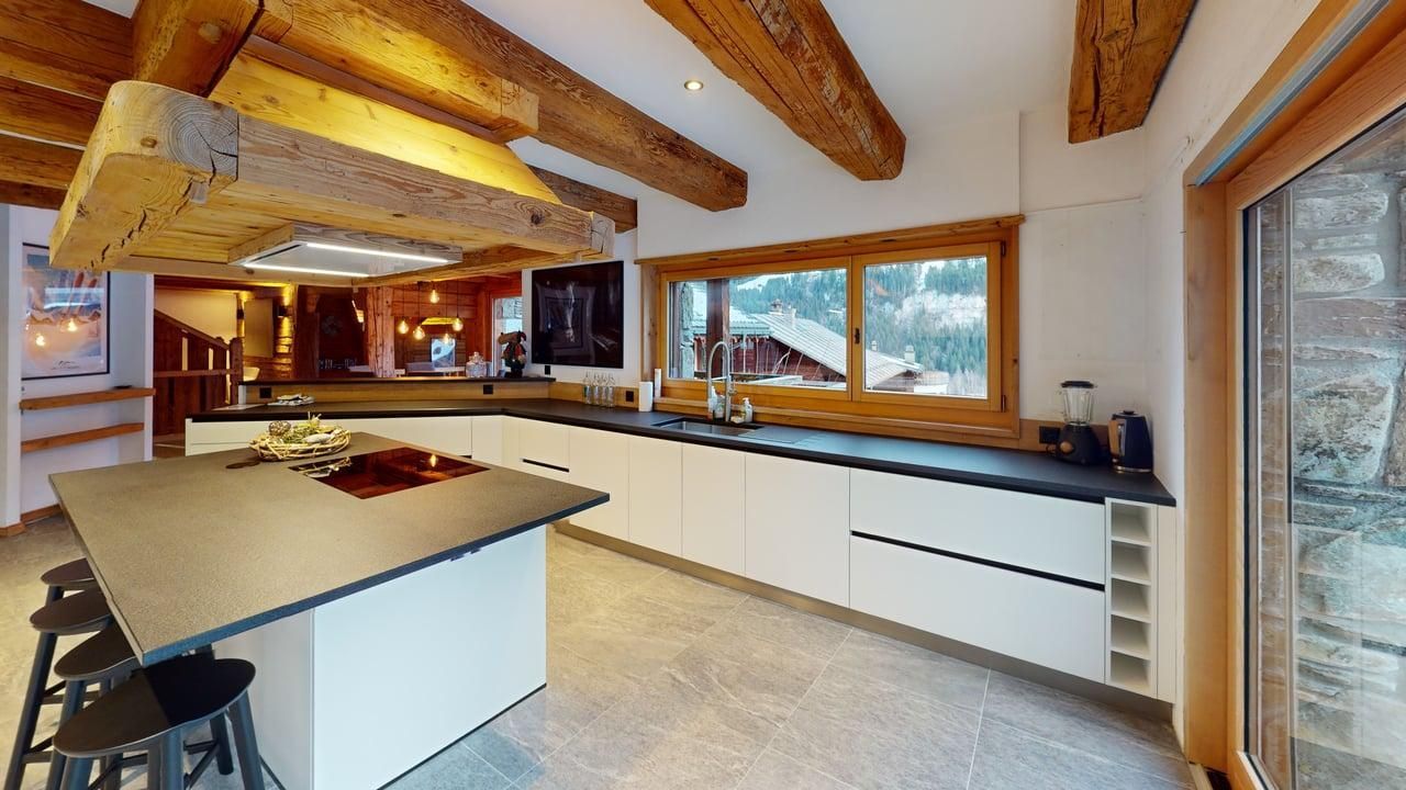 A bright modern kitchen with white cabinets, dark stone countertops, an island with bar stools, and rustic wood beams.
