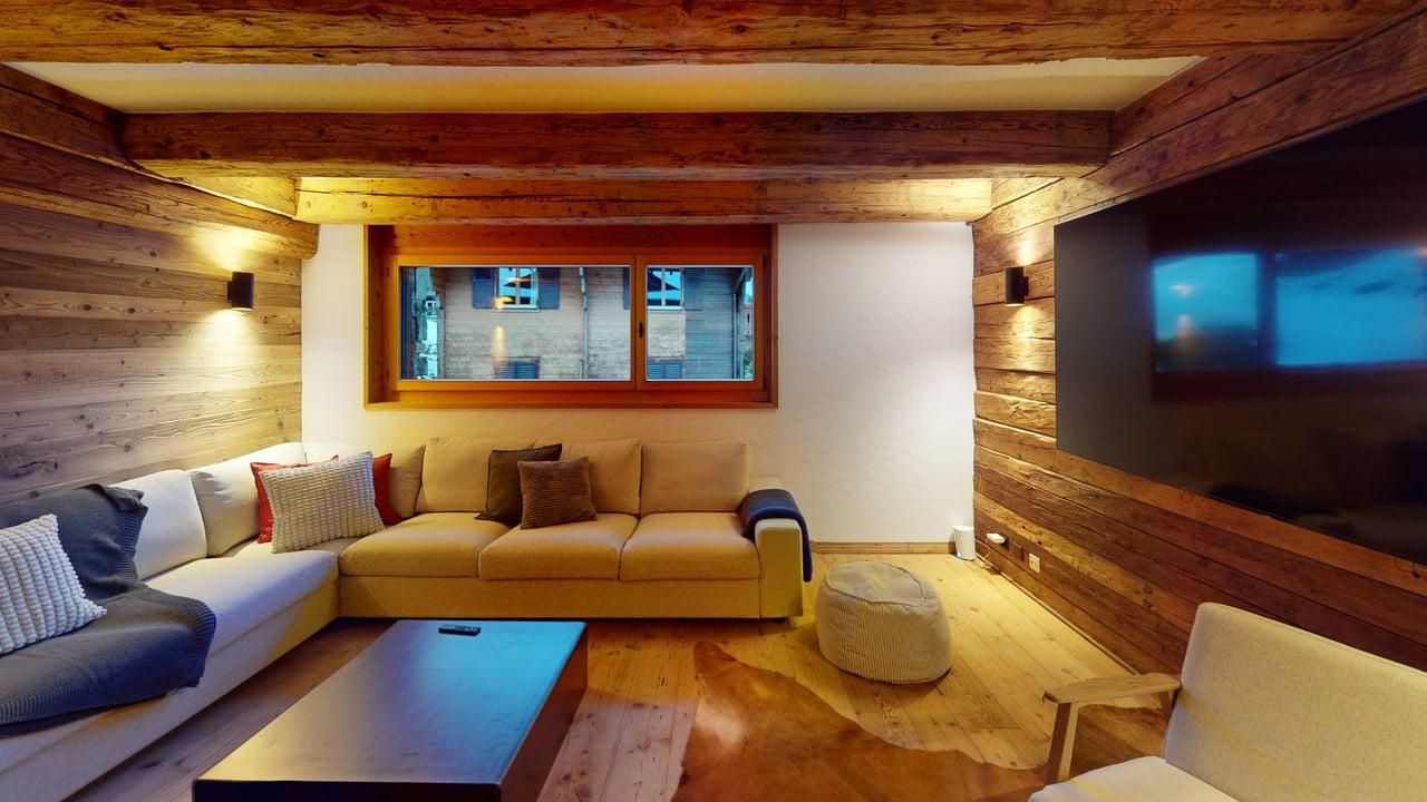 A cozy living room with wooden walls, ceiling beams, a large sofa, a coffee table, and a wall-mounted television.