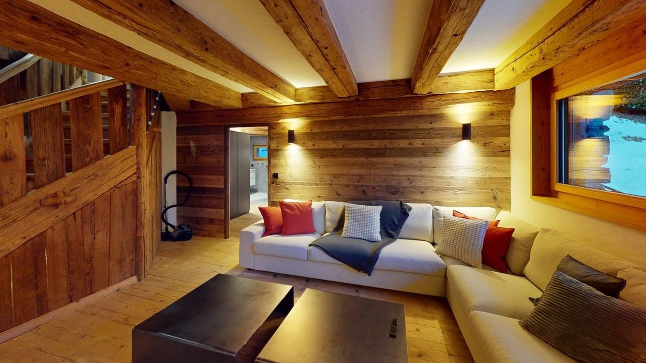 A cozy rustic living room with wooden walls and ceiling beams, featuring a large sectional sofa and two dark coffee tables.