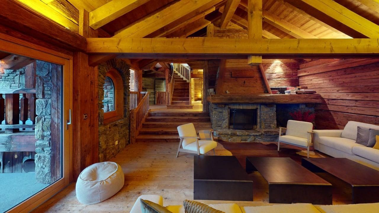 A rustic, spacious living room with wooden walls and ceiling, stone fireplace, comfortable seating, and indoor stairs.