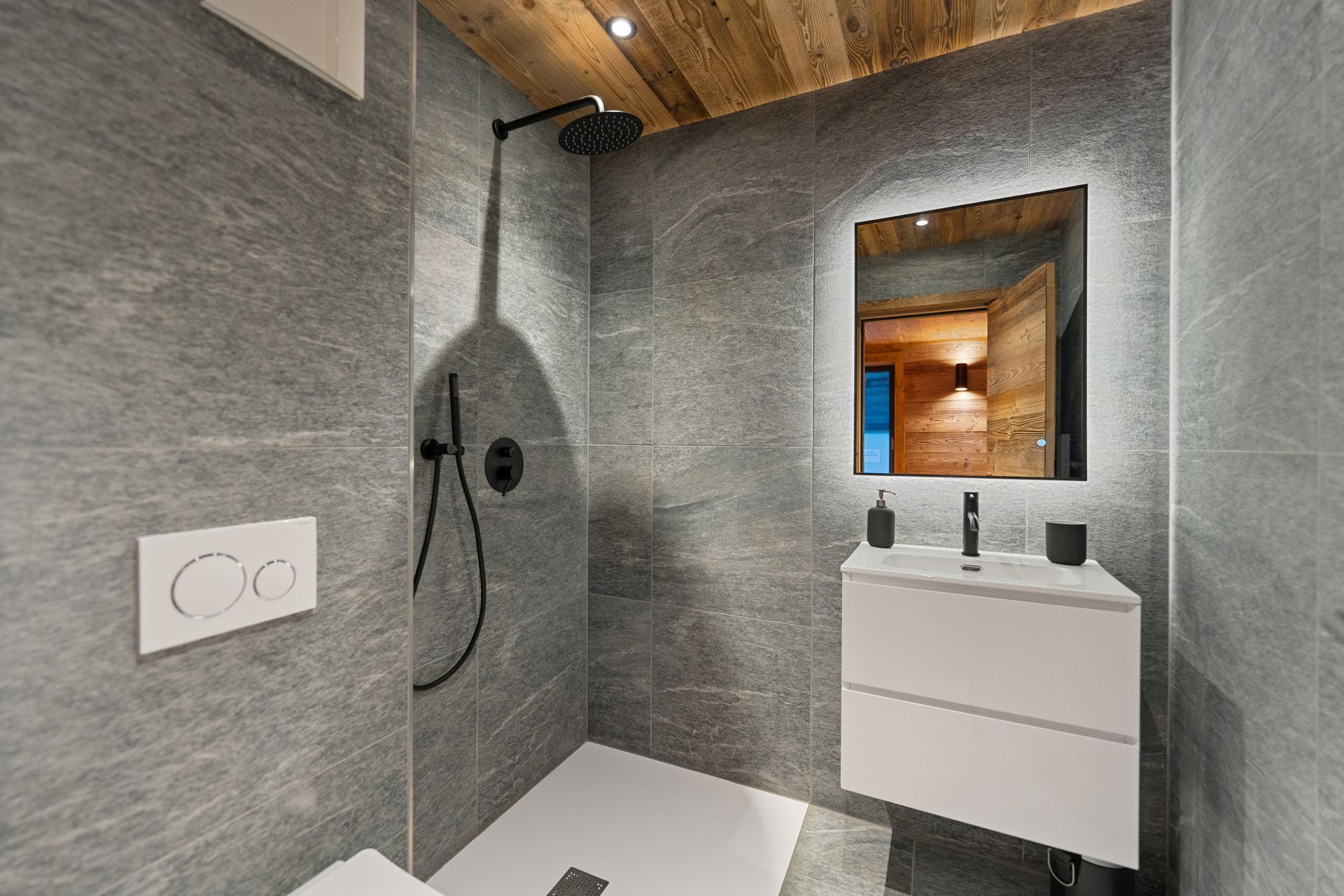 Modern bathroom with gray stone tiles, a white wall-mounted vanity, a backlit mirror, and a walk-in shower.