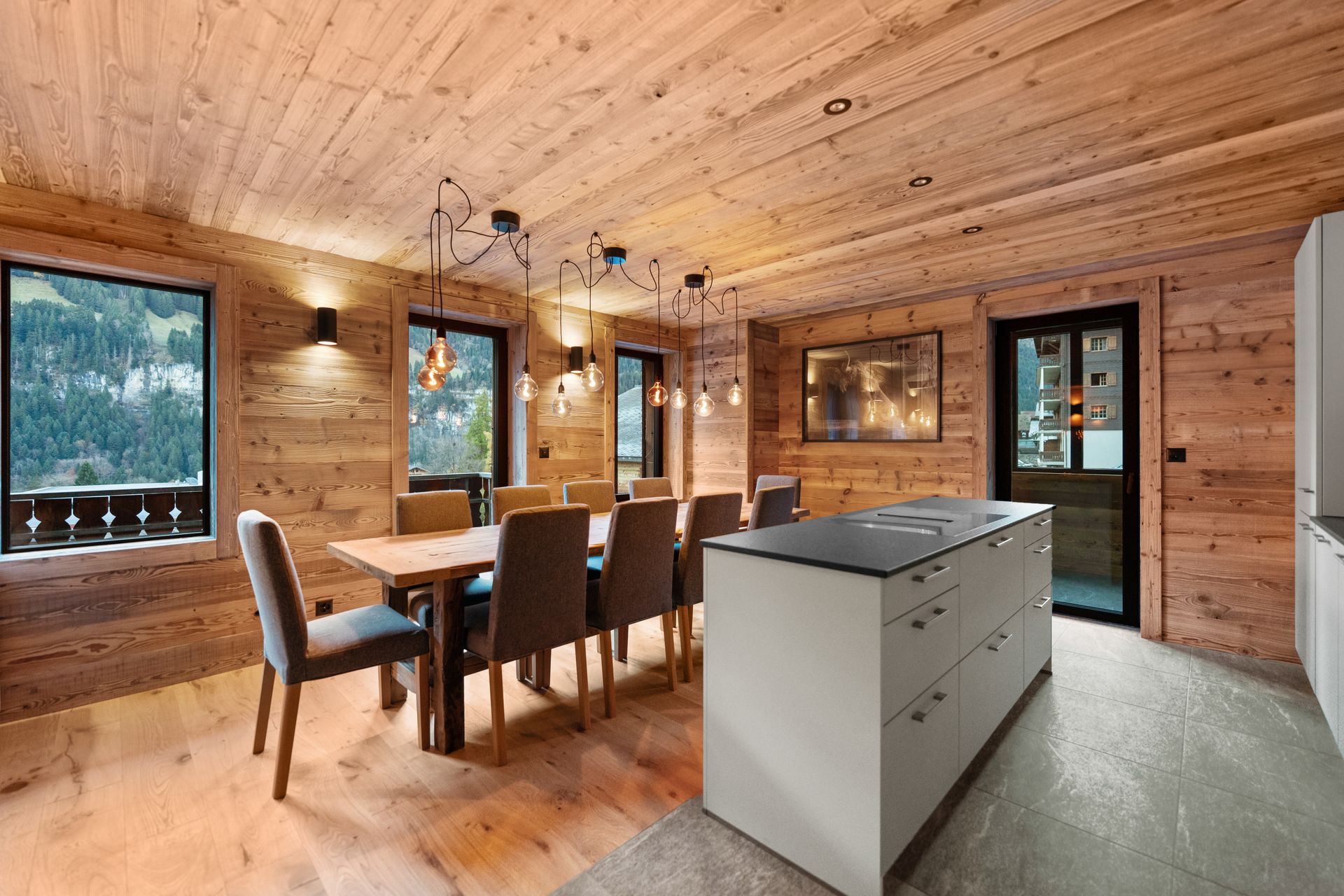 A modern wooden dining room with a long table, grey chairs, pendant lights, and a kitchen island with a stone countertop.