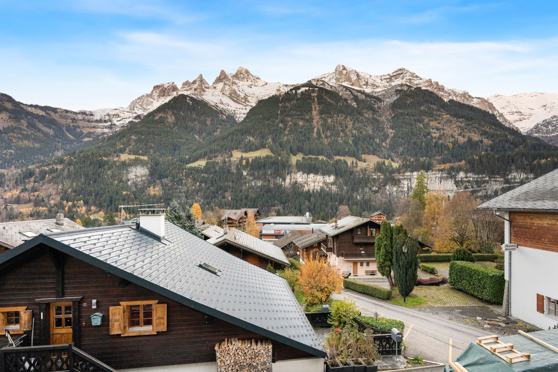 A dark wooden cabin with a grey roof overlooks a mountain village with snow-capped peaks in the background.