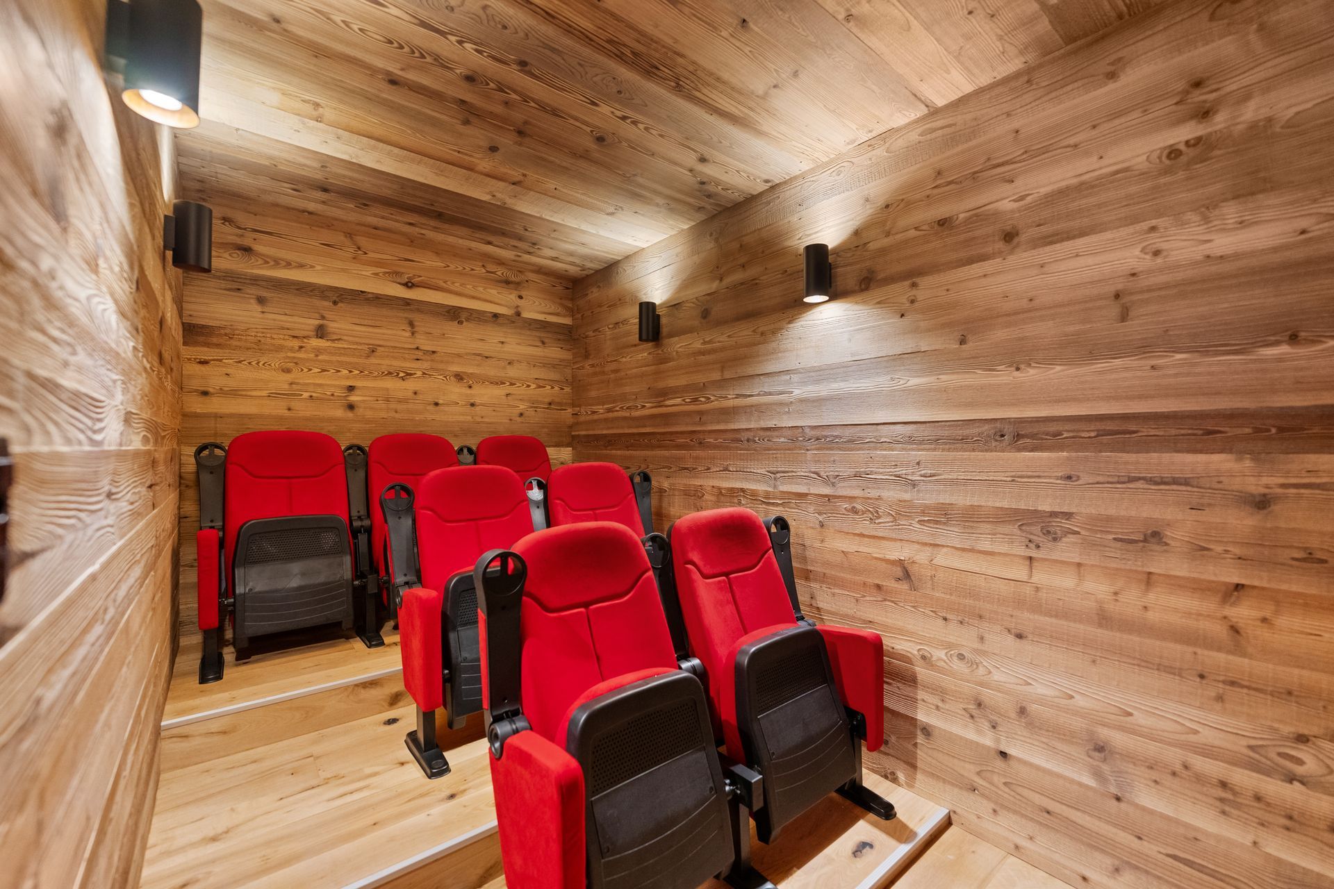 A small home cinema with two rows of red seats on a raised platform, set in a room with light-toned wooden walls.
