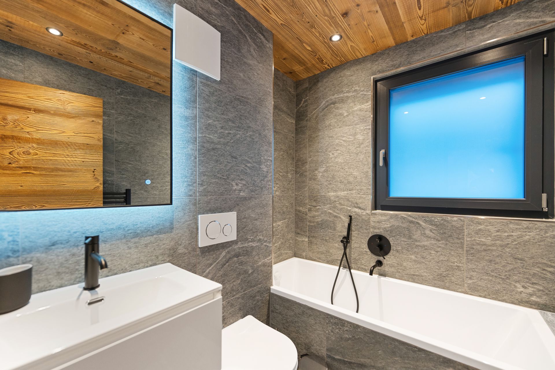 A modern bathroom featuring stone walls, a wooden ceiling, a white vanity, a toilet, and a bathtub under a frosted window.