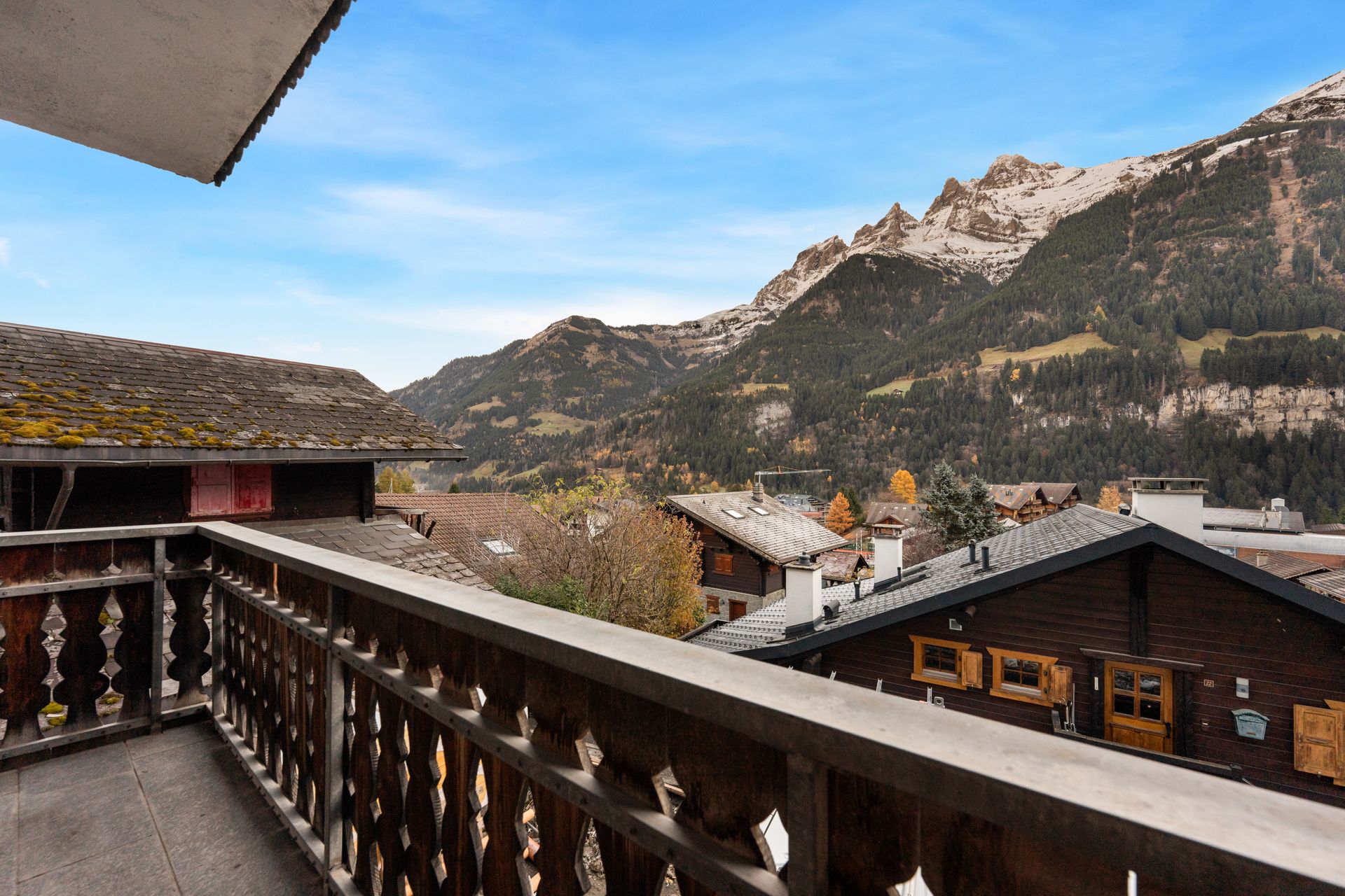 A wooden balcony overlooking a village in a valley with snow-capped mountains under a clear blue sky.