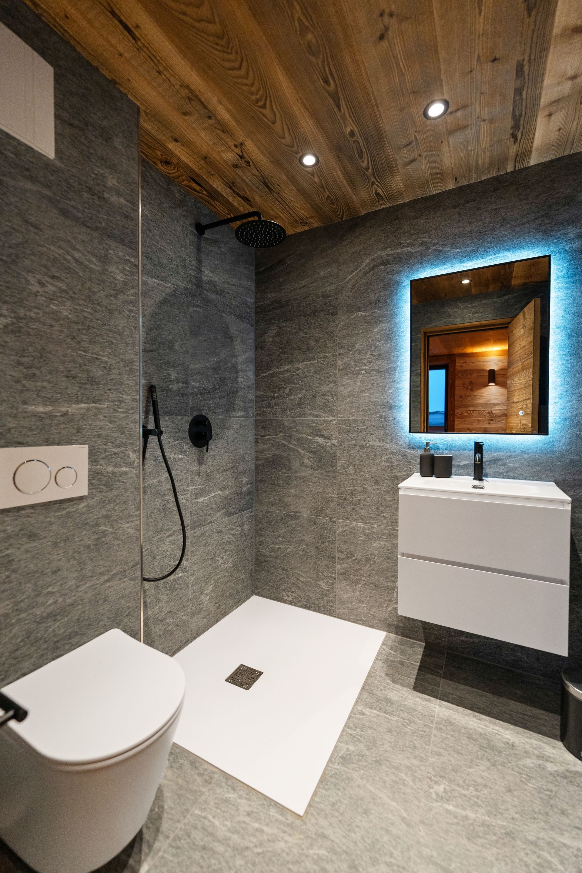 Modern bathroom with gray stone walls, a wooden ceiling, walk-in shower, wall-mounted toilet, and illuminated vanity mirror.