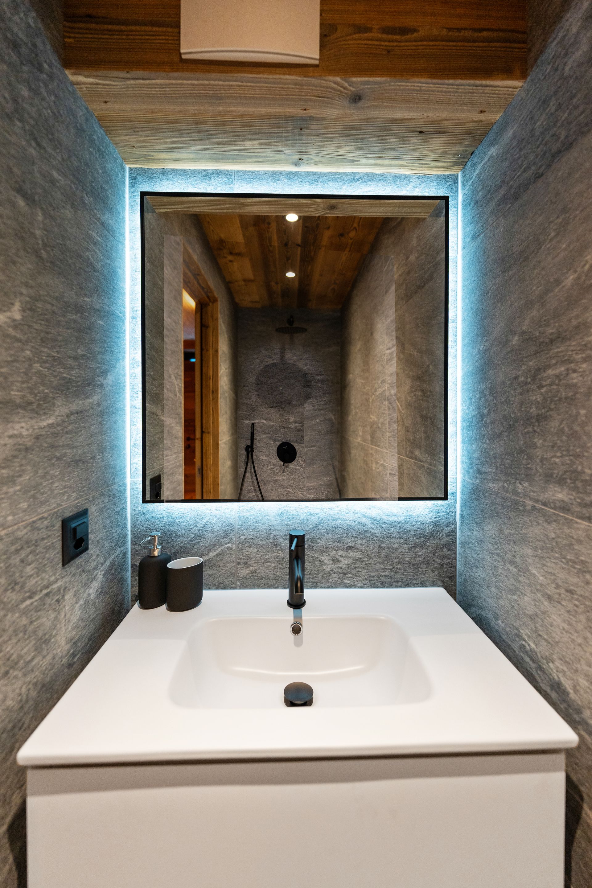 A modern bathroom vanity with a white basin and black faucet, featuring a backlit mirror against gray stone walls.