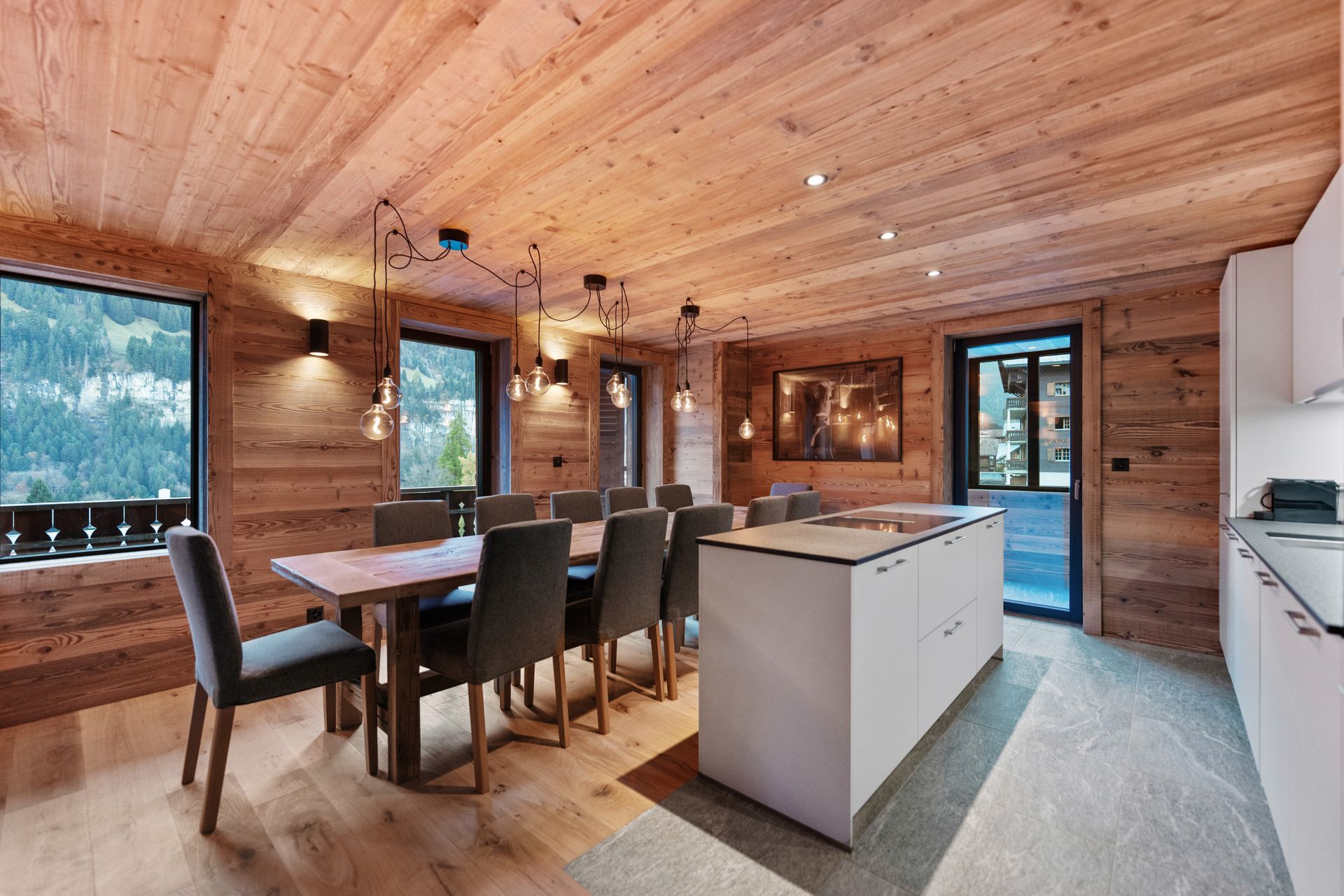 A modern kitchen and dining area in a wooden chalet featuring a white island, wooden table with gray chairs, and windows.