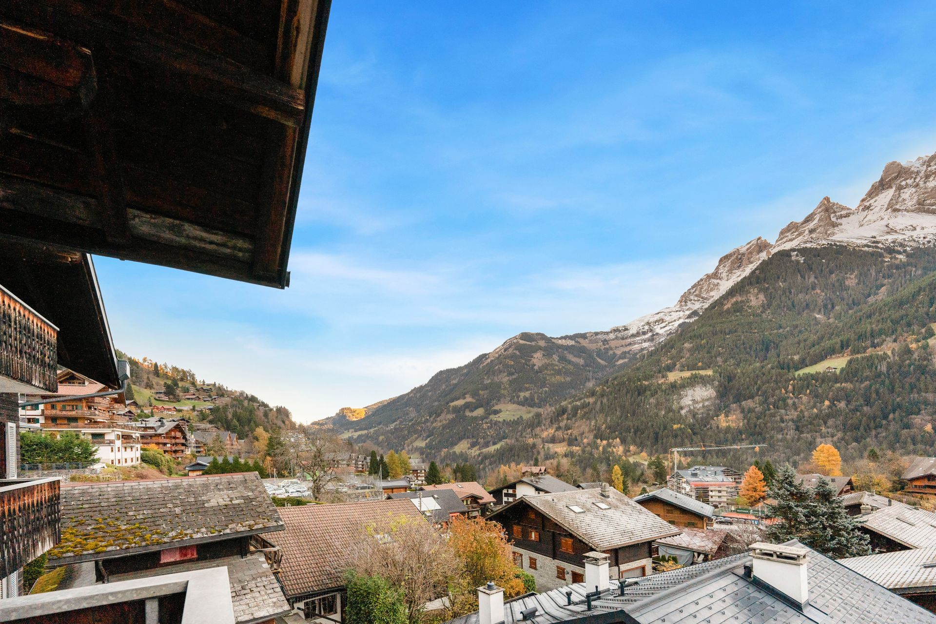Vue panoramique d'un village de montagne avec ses chalets en bois, ses arbres luxuriants et ses sommets imposants saupoudrés de neige sous un ciel bleu.