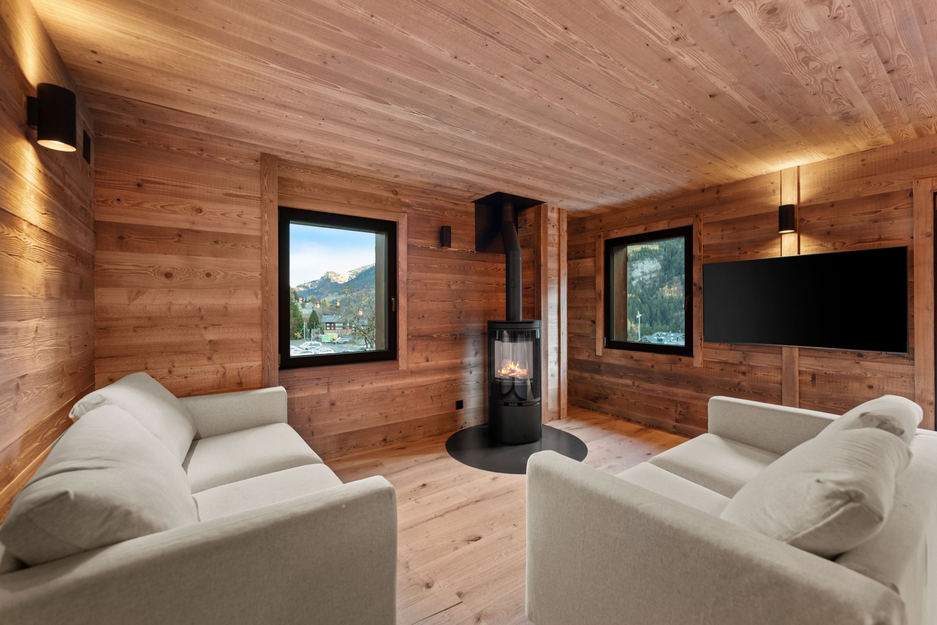 A modern living room with wood paneled walls, two light-colored sofas facing a fireplace, and a wall-mounted TV.