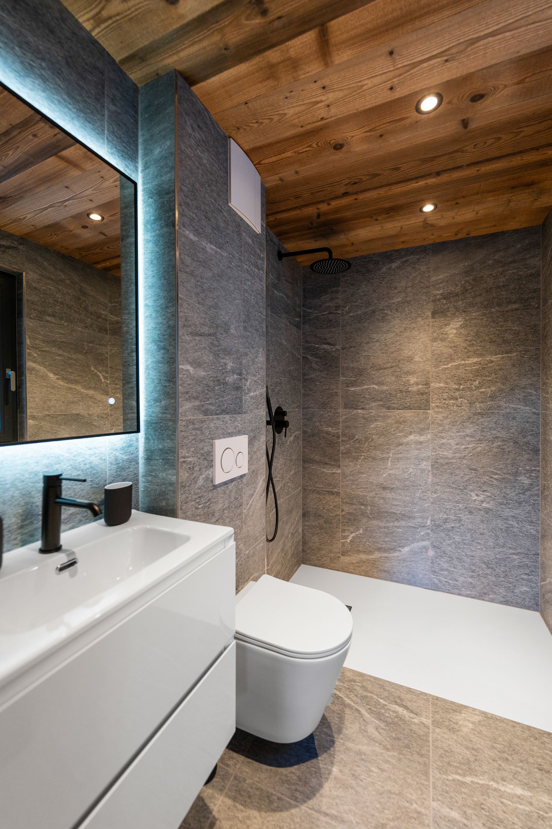 Une salle de bains moderne avec un plafond lambrissé, des murs en pierre grise, des toilettes suspendues blanches et un lavabo blanc.