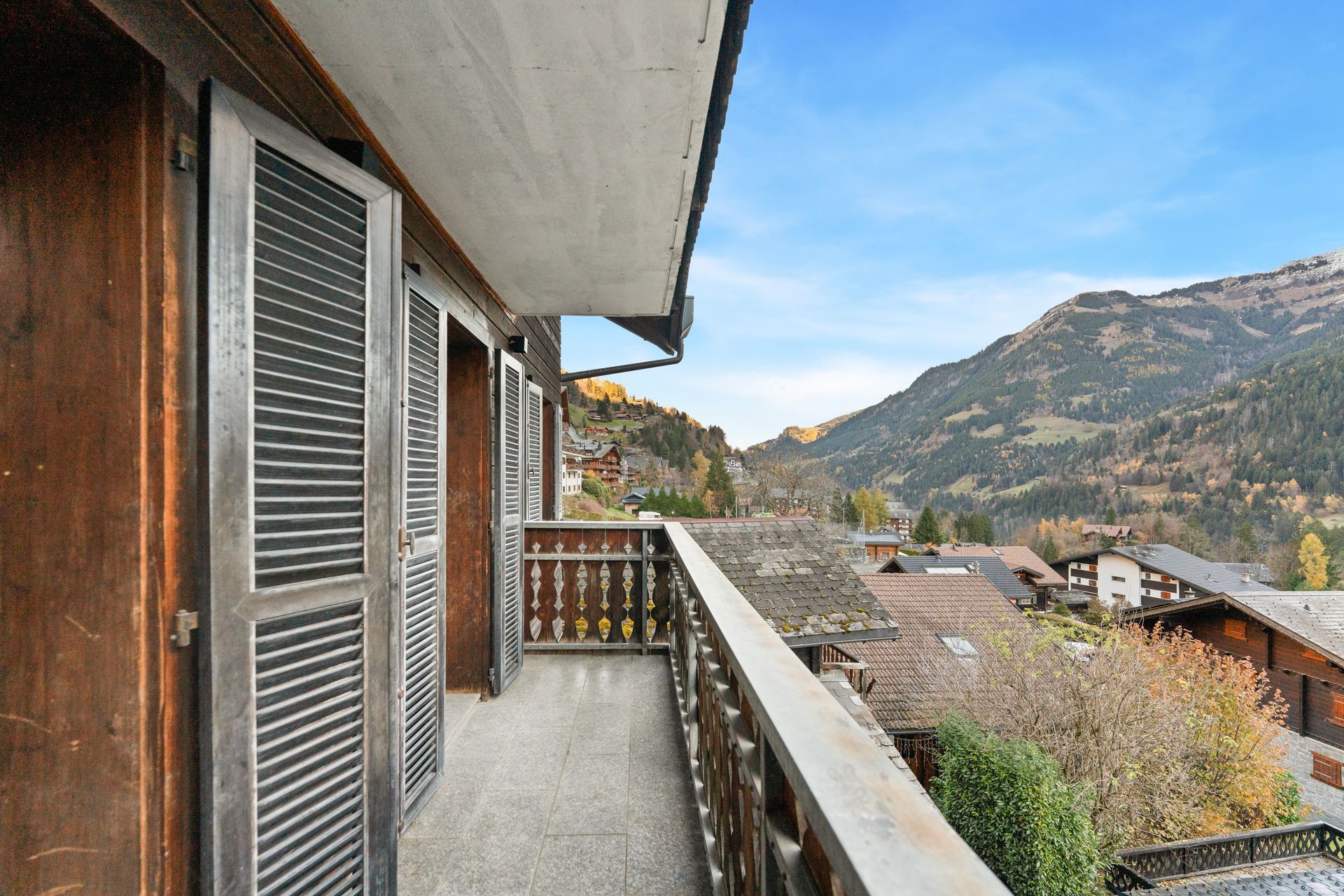 Un balcon avec des volets en bois et une rambarde surplombe un pittoresque village de montagne sous un ciel bleu.