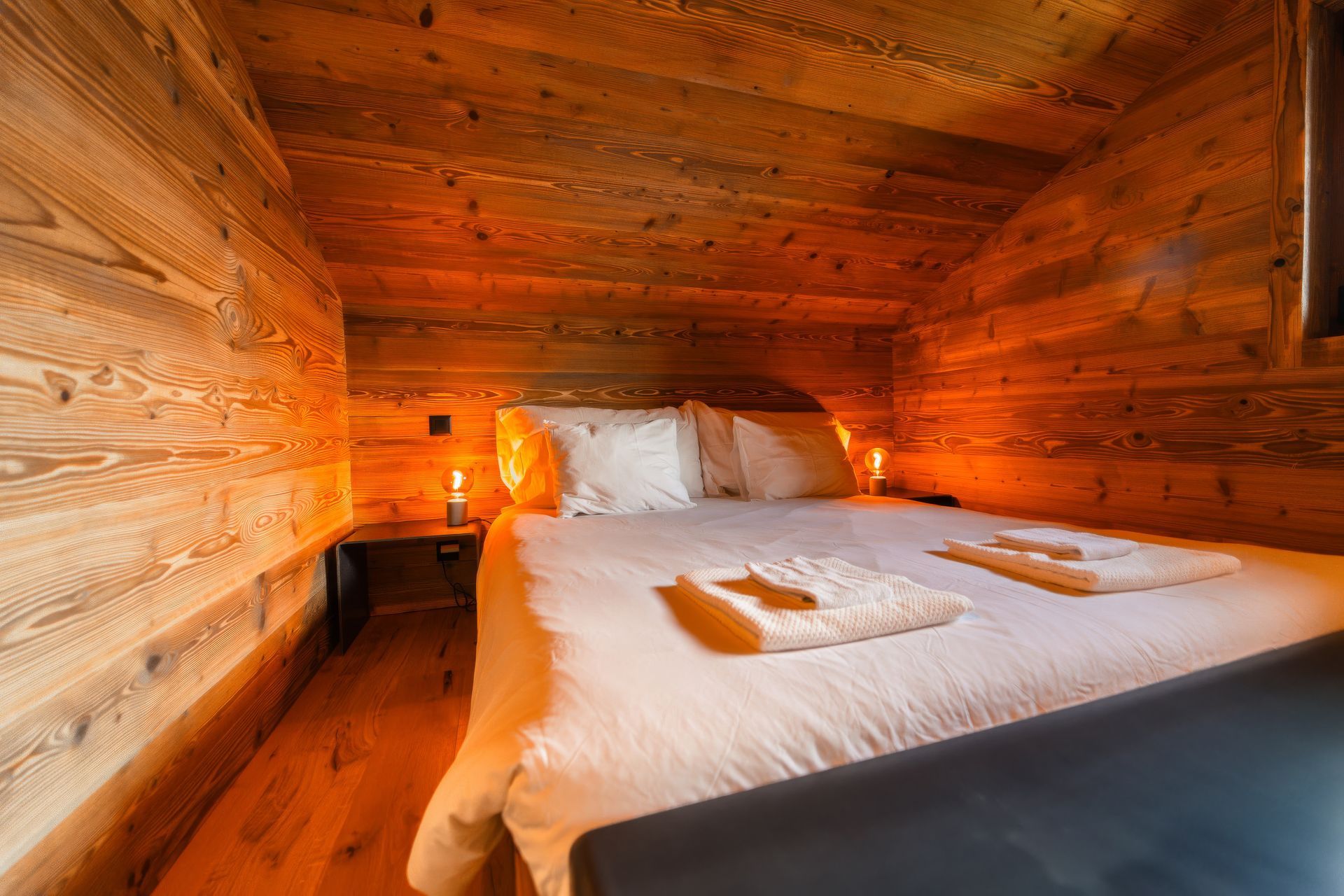A cozy bedroom with a wooden-paneled slanted ceiling, a bed with white linens, and warm, soft lighting.