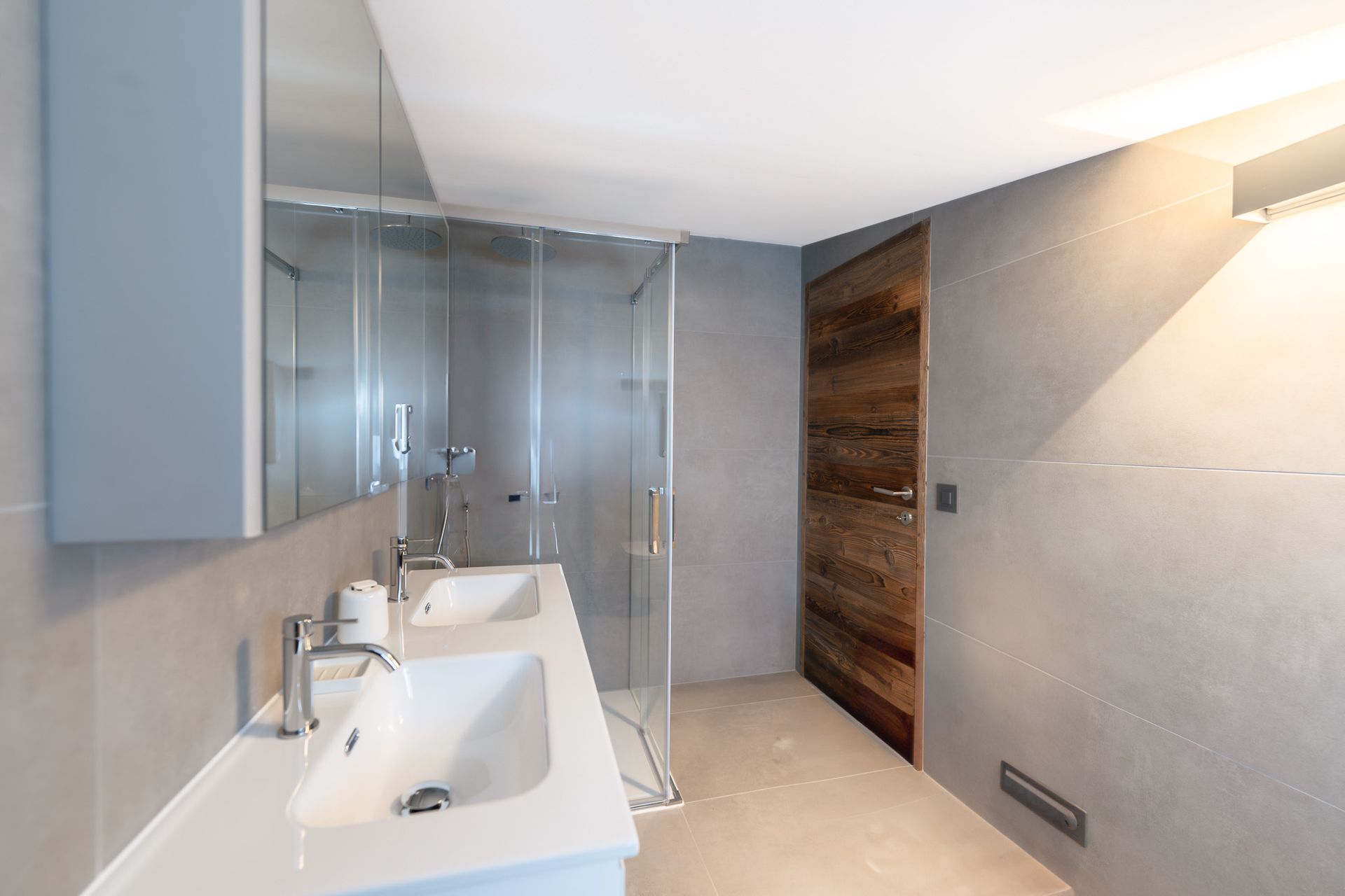 A modern bathroom with a double white vanity, a glass-enclosed shower, gray tile walls, and a rustic wooden door.