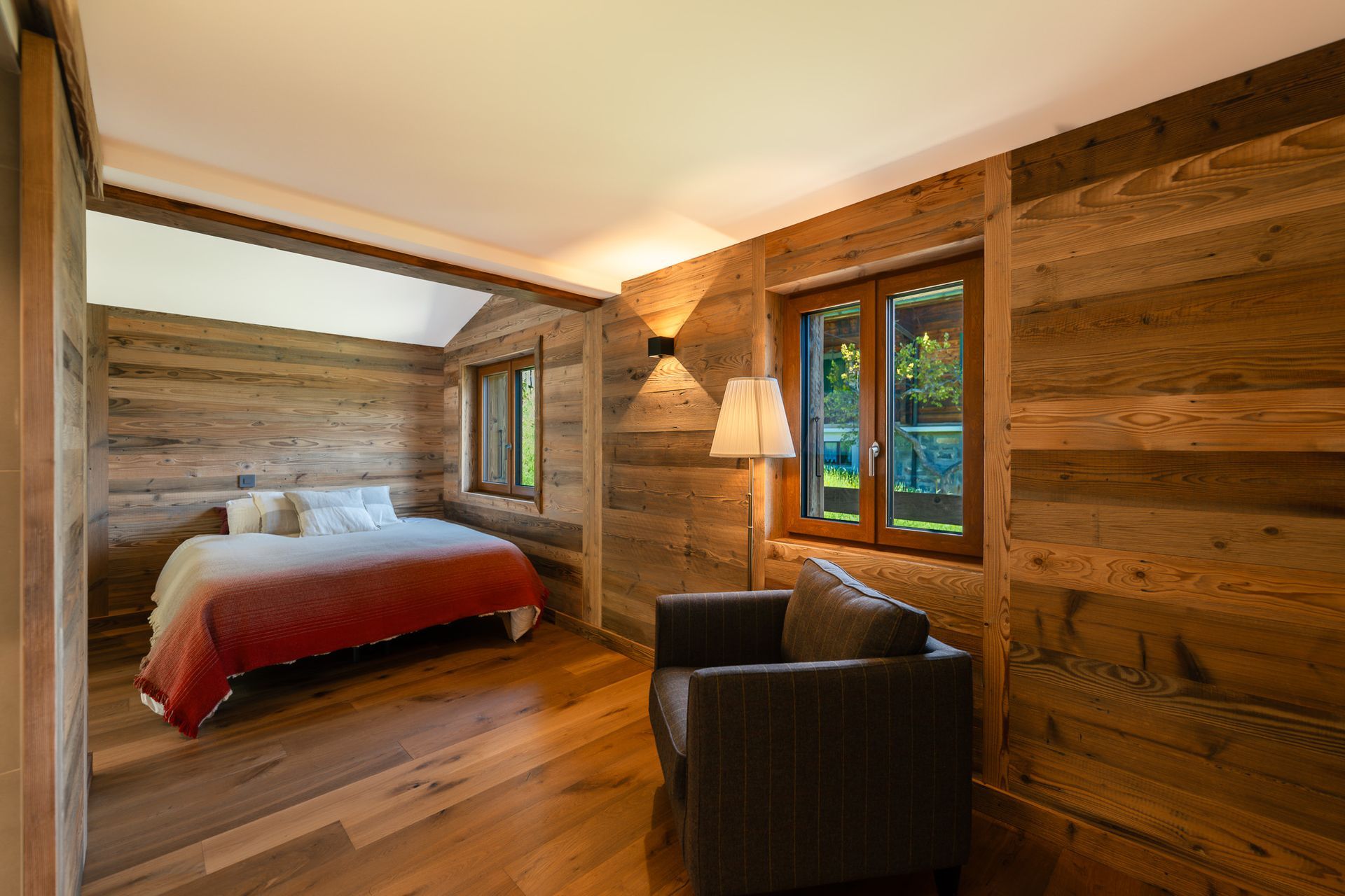 A rustic bedroom with wood-paneled walls, a bed featuring a red and white blanket, and a dark armchair near a window.