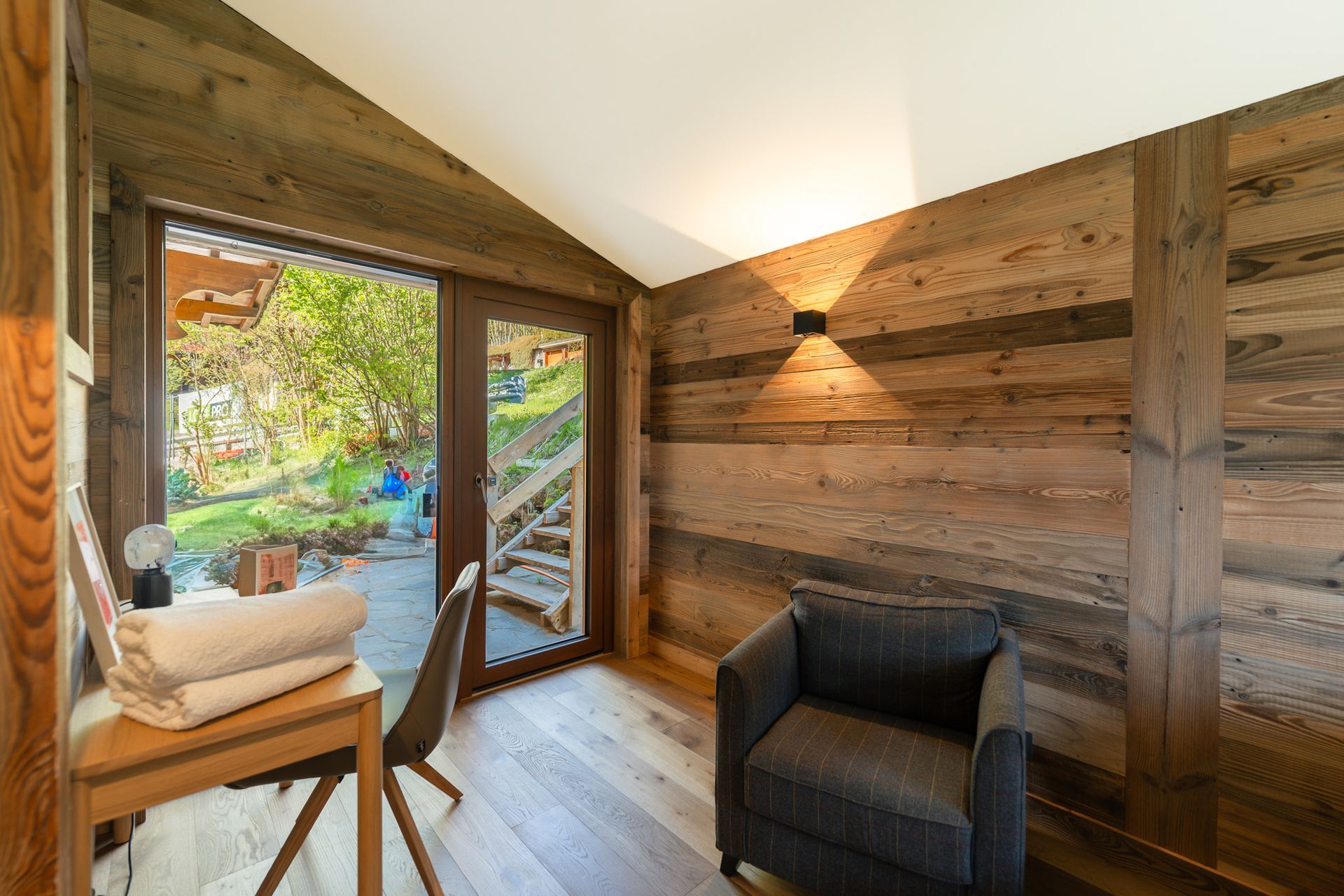 A cozy room with wood-paneled walls, an armchair, a desk with folded towels, and a glass door opening to a garden.