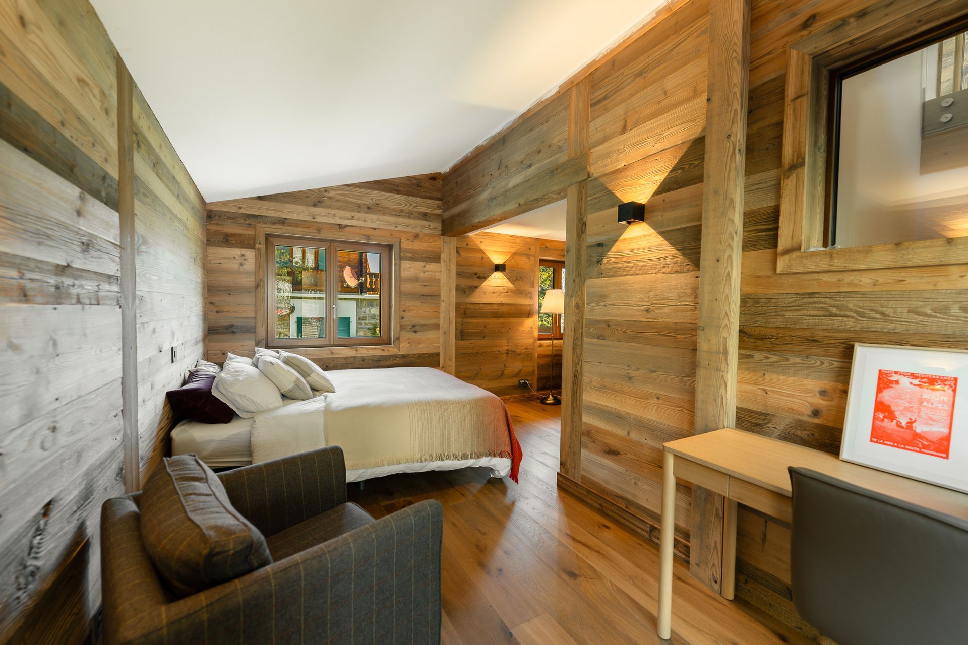 A rustic bedroom with wooden walls, a neatly made bed, an armchair, and a small desk with framed artwork.