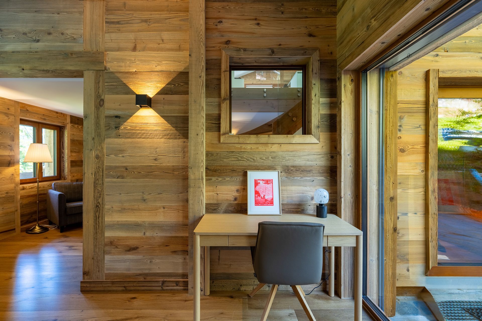 A wooden room with a desk, grey chair, and desk lamp, featuring an adjacent seating area viewed through an open doorway.