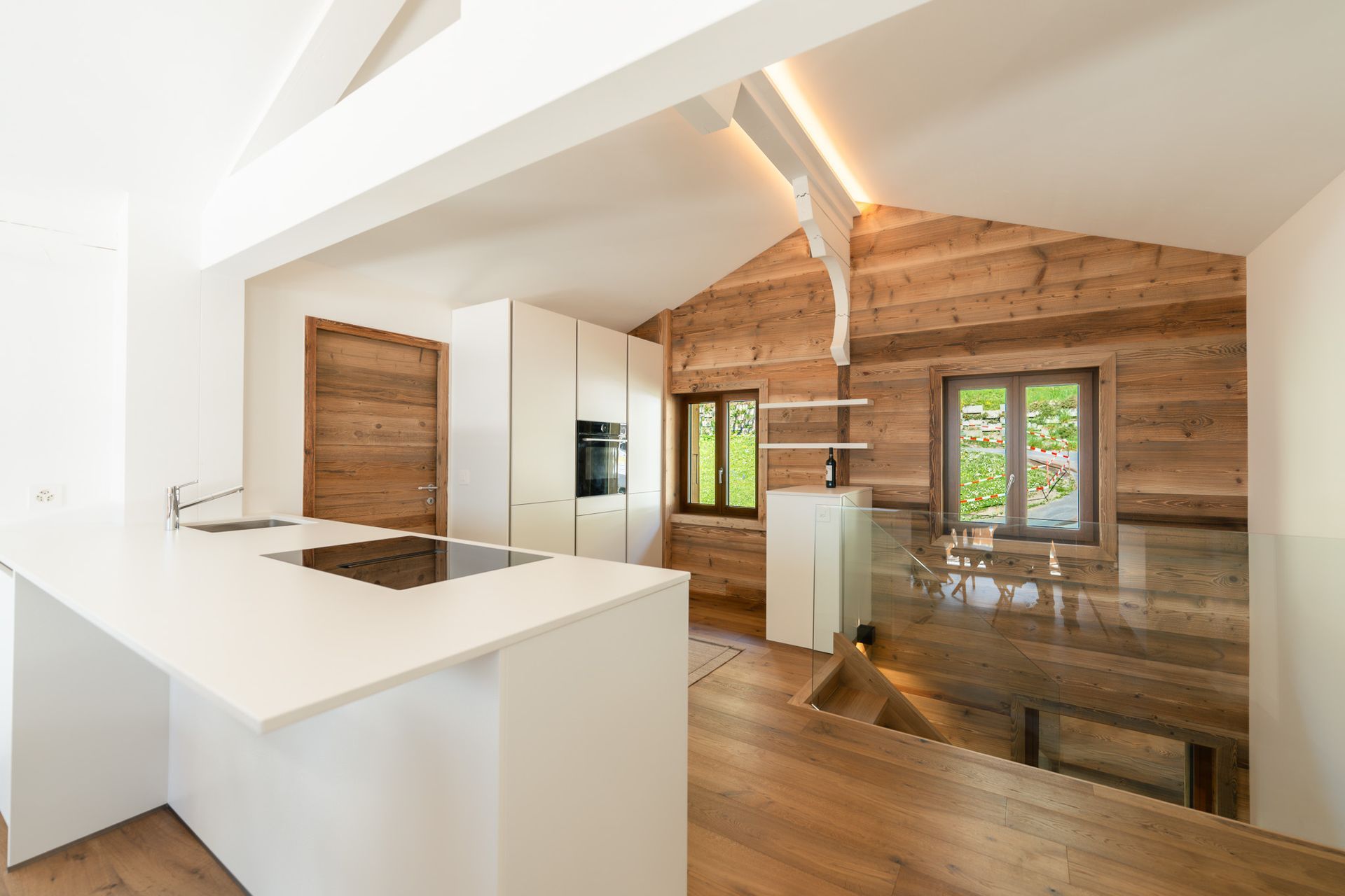 A bright, modern kitchen with a white island and cabinets contrasted against rustic wooden walls, featuring a glass rail.