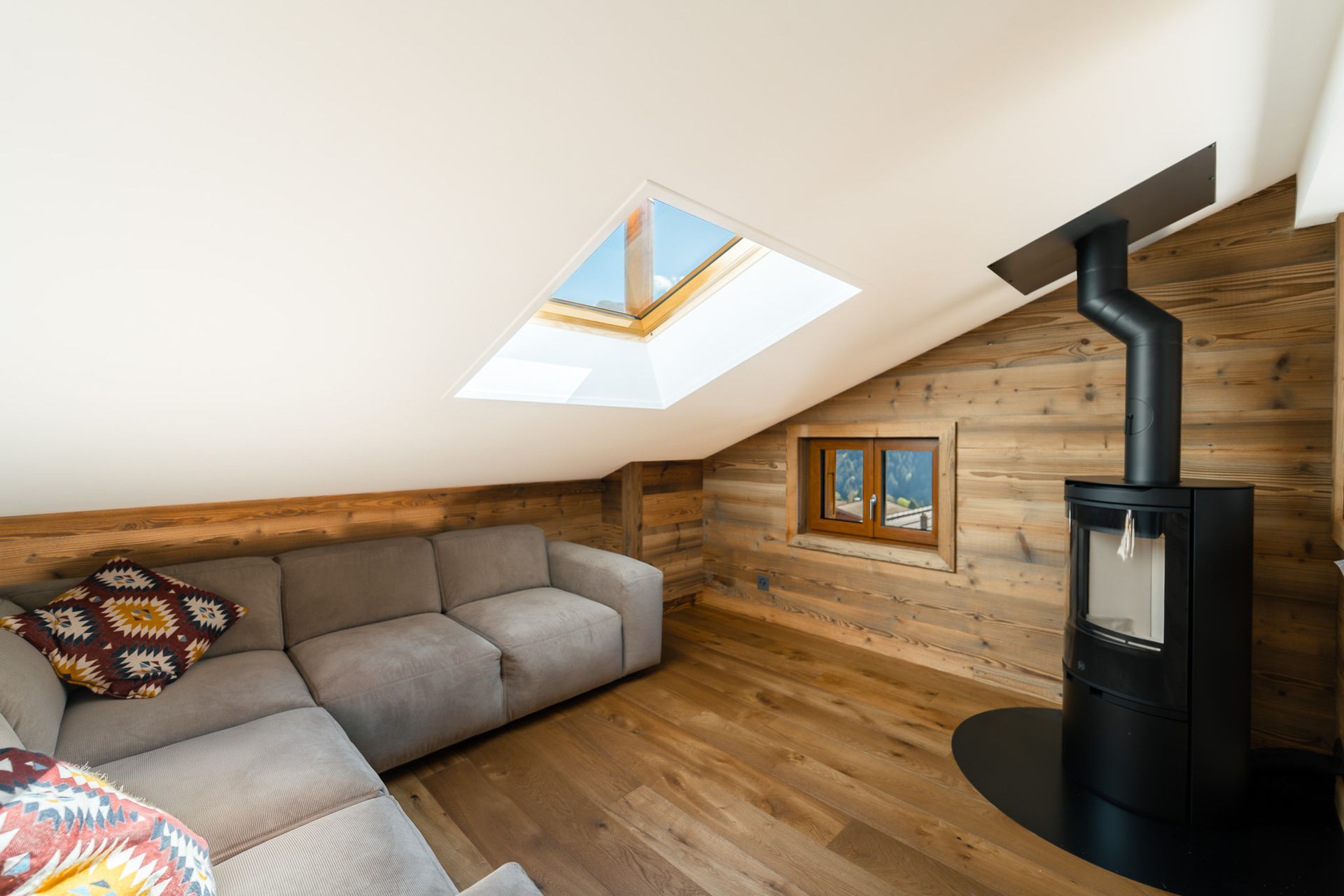 A cozy attic living room with light wood walls, a grey sectional sofa, a skylight, and a black wood-burning stove.