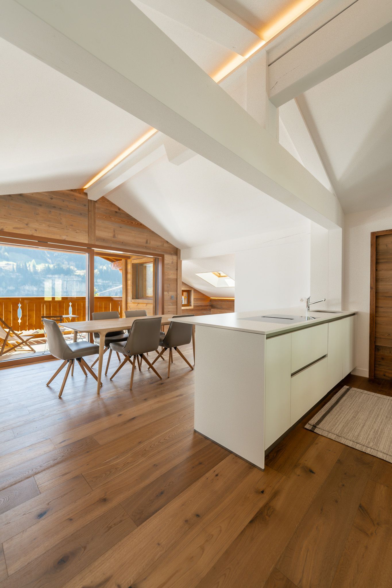 A modern, bright kitchen area with white cabinets and a dining table, featuring wooden floors and mountain views.