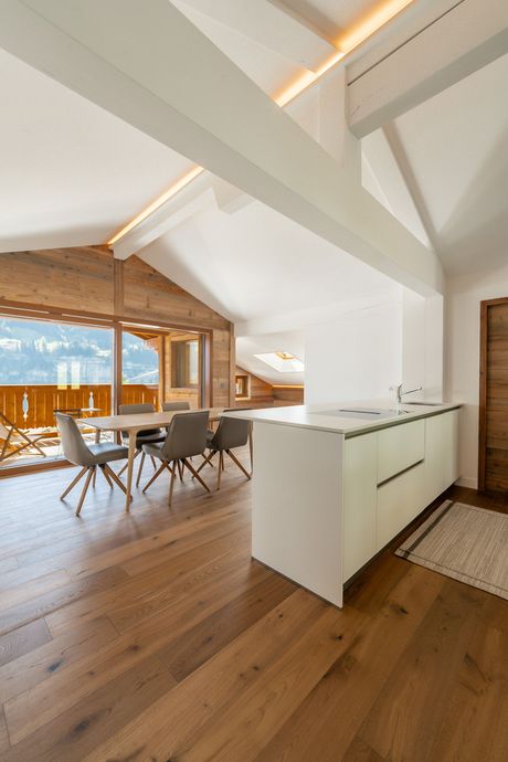 Cuisine et salle à manger modernes dans une maison de montagne avec parquet, meubles blancs et grandes fenêtres donnant sur un balcon.