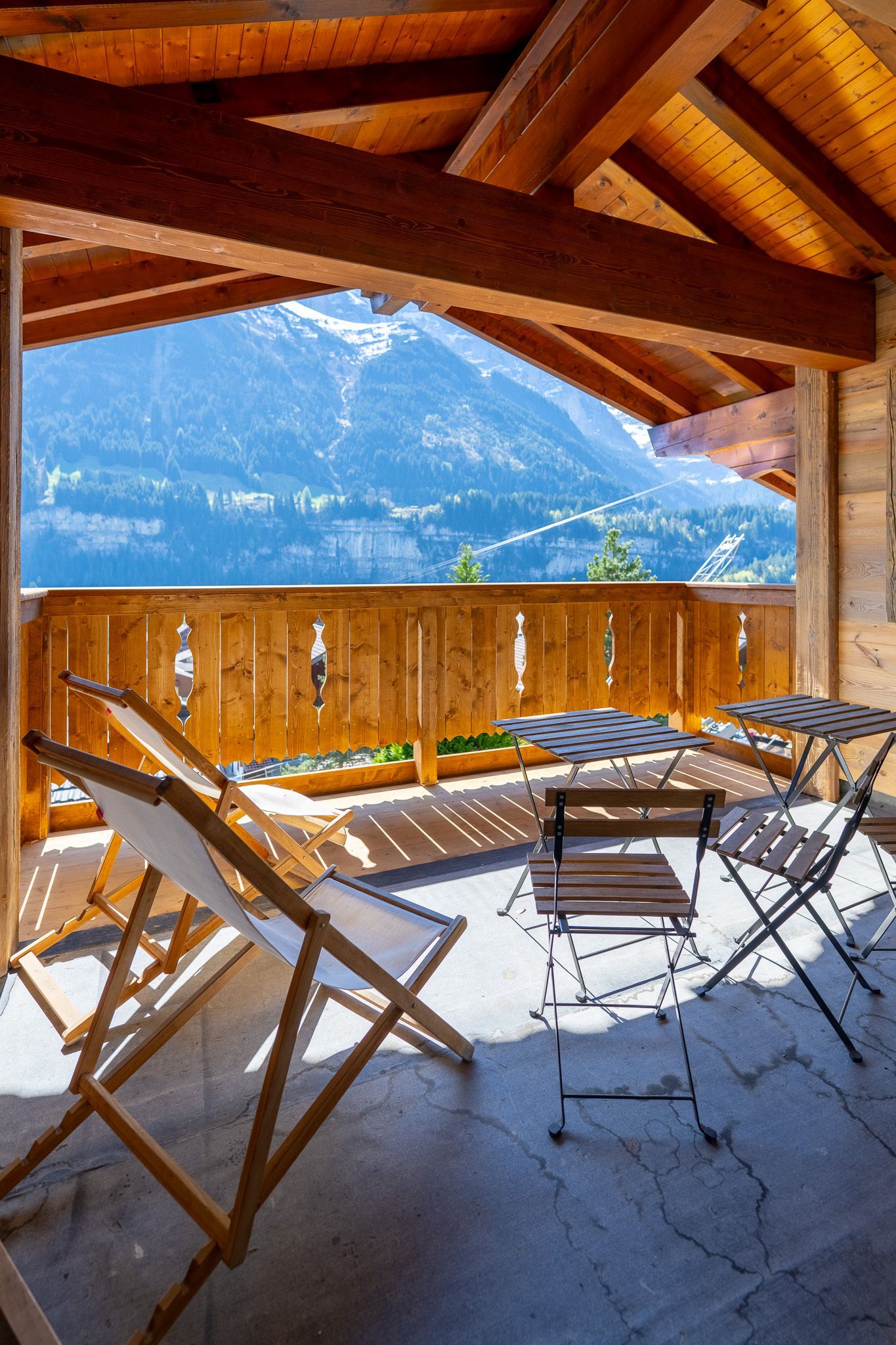A balcony with wooden deck chairs and tables overlooking a scenic, snow-capped mountain range under a clear blue sky.