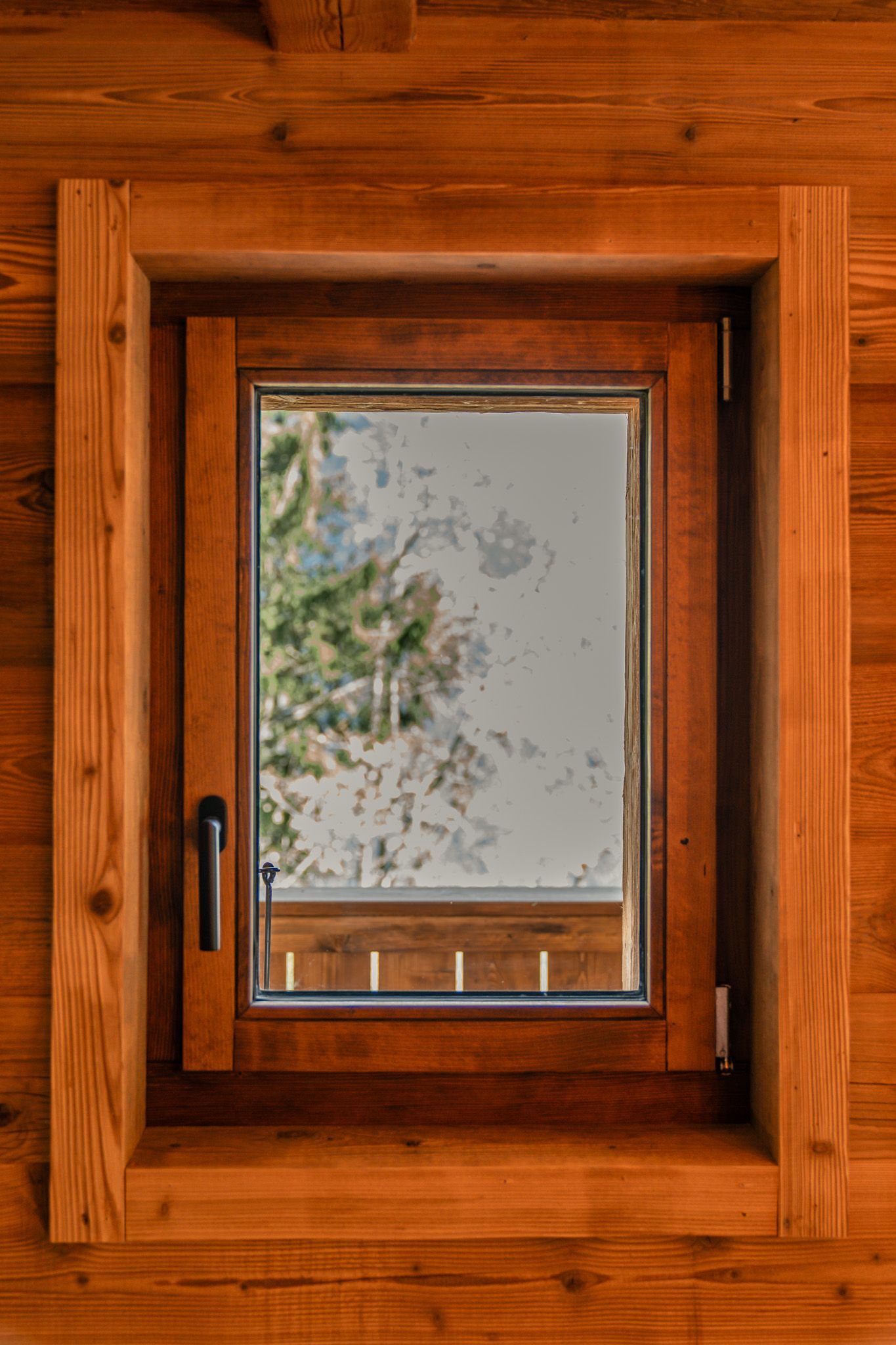 A view of a snowy landscape through a wooden-framed casement window set into a rustic log cabin wall.