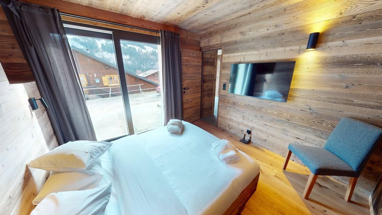Une chambre confortable aux murs lambrissés, avec un grand lit, une télévision murale et un fauteuil bleu près d'une porte-fenêtre donnant sur un balcon avec vue.