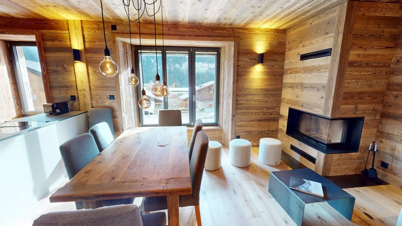 Une salle à manger rustique avec des murs en bois, une grande table, des chaises grises, des tabourets et une cheminée d'angle près d'une fenêtre.